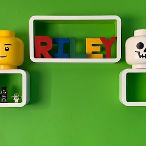 Może przedstawiać: Zielona ściana z białymi p&oacute;łkami z dekoracjami w stylu Lego. Słowo "RILEY" jest zapisane kolorowymi klockami Lego. Ż&oacute;łta głowa Lego i głowa Lego z czaszką są eksponowane na p&oacute;łkach, wraz z figurkami Lego.