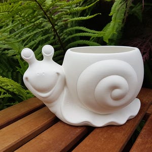 May include: A matte white ceramic snail-shaped planter featuring a smooth, rounded shell body that serves as a container, a smiling face with two protruding eye stalks, and a wide, flat base designed to sit securely on a flat surface.