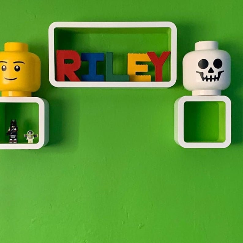 Może przedstawiać: Zielona ściana z białymi p&oacute;łkami z dekoracjami w stylu Lego. Słowo "RILEY" jest zapisane kolorowymi klockami Lego. Ż&oacute;łta głowa Lego i głowa Lego z czaszką są eksponowane na p&oacute;łkach, wraz z figurkami Lego.