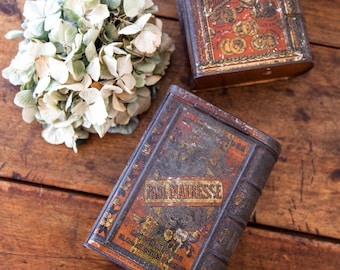 May include: Two vintage metal tins with book-shaped designs. The top tin is red and gold with the text "Chocolat Française" and "Paris Delices". The bottom tin is brown and gold with the text "Paul Mairesse" and "Maison Paris".