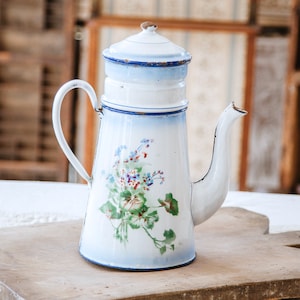Puede incluir: Cafetera de esmalte blanco con ribete azul y diseño floral. La cafetera tiene tapa y pico. Está sobre una tabla de cortar de madera.