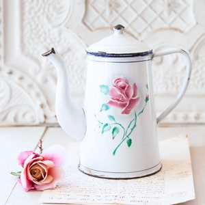 May include: White enamelware coffee pot with a pink rose design. The pot has a long spout, a handle, and a lid. A pink rose and a piece of aged paper are next to the pot.