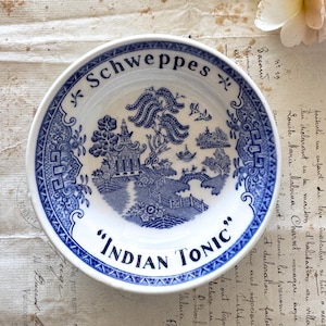 May include: A small, round, white and blue ceramic bowl with the text "Schweppes" and "Indian Tonic". The bowl features a blue and white landscape design. A rose and stem are on the background.