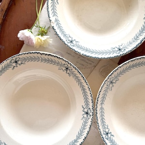 May include: A collection of off-white ceramic bowls with a blue leaf and ribbon border design. The bowls are arranged on a wooden surface, with a few flowers and vintage paper visible in the background. The bowls are of varying sizes.
