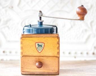 May include: Vintage wooden coffee grinder with a blue metal top and a wooden handle. The grinder has a drawer and a label that reads "Peugeot Paris".