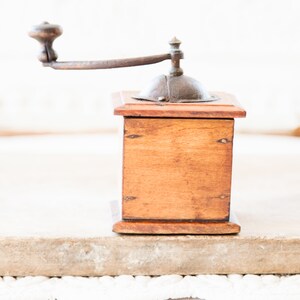 May include: A vintage wooden coffee grinder with a metal crank and top. The wooden box is a warm brown color, and the metal components have a rustic, aged appearance. The grinder is designed for manual operation.