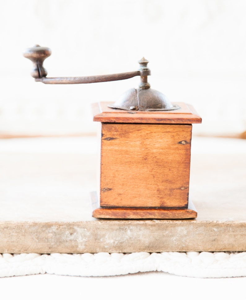 May include: A vintage wooden coffee grinder with a metal crank and top. The wooden box is a warm brown color, and the metal components have a rustic, aged appearance. The grinder is designed for manual operation.
