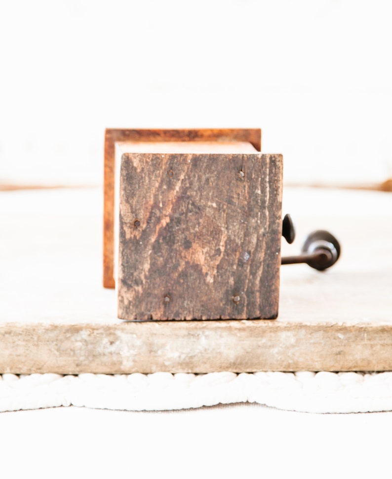 May include: A vintage wooden coffee grinder with a dark, weathered appearance. The grinder features a square wooden body with visible wood grain and a metal handle. The design suggests a rustic, antique aesthetic.