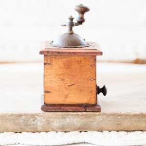 May include: A vintage wooden coffee grinder with a metal top and crank. The wooden box is a warm brown color, and the metal components show signs of age. The grinder is a classic kitchen tool.