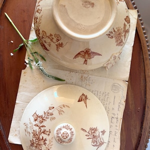 Puede incluir: Cuenco de cerámica antigua con tapa, con motivos florales y de pájaros marrones. El cuenco y la tapa son de color blanquecino con un acabado brillante. La tapa tiene un pomo decorativo. El cuenco está sobre una mesa de madera.