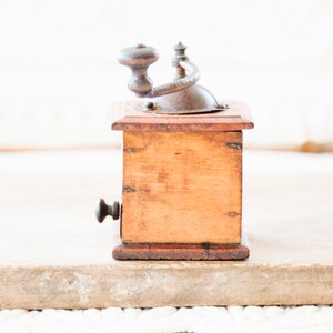 May include: A vintage wooden coffee grinder with a metal crank and handle. The wooden body is a warm brown color, and the metal components show signs of age. The grinder is set against a neutral background, highlighting its classic design.
