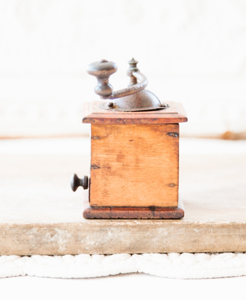 May include: A vintage wooden coffee grinder with a metal crank and handle. The wooden body is a warm brown color, and the metal components show signs of age. The grinder is set against a neutral background, highlighting its classic design.