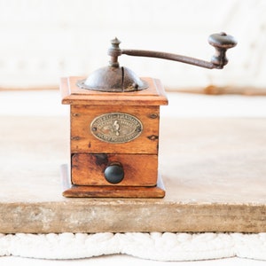 May include: A vintage wooden coffee grinder with a metal crank and a small drawer. The grinder has a dark brown finish and a metal plate with text. The handle is dark brown and the drawer has a black knob.