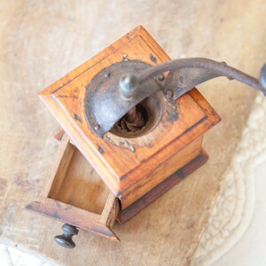 May include: A vintage wooden coffee grinder with a metal crank and a small drawer. The grinder is made of light brown wood and has a dark metal top. The drawer is open, revealing the interior. The grinder sits on a textured surface.