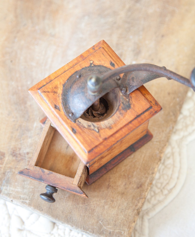 May include: A vintage wooden coffee grinder with a metal crank and a small drawer. The grinder is made of light brown wood and has a dark metal top. The drawer is open, revealing the interior. The grinder sits on a textured surface.