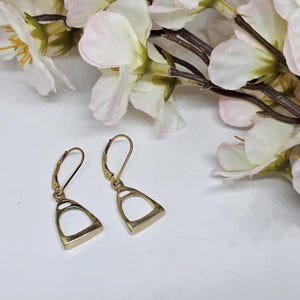 Puede incluir: Un par de pendientes dorados con forma de estribo y cierre de palanca. Los pendientes se exhiben sobre una superficie blanca, con flores blancas y rosas en el fondo. Los pendientes tienen un diseño clásico inspirado en la equitación.