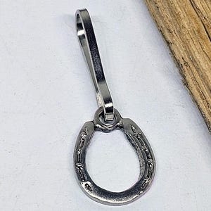 May include: A silver-colored horseshoe keychain with a long, curved clip. The horseshoe has detailed markings and is connected to the clip by a small ring. The keychain is made of metal.