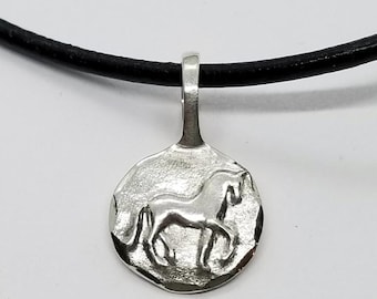 May include: A silver-colored circular pendant featuring a raised relief of a horse in profile. The pendant has a textured, uneven surface and is attached to a simple, smooth black cord necklace that runs horizontally across the top of the frame.