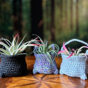 May include: Three crocheted plant pots in different colors, black, blue and purple, with air plants inside. The pots are sitting on a wooden table.