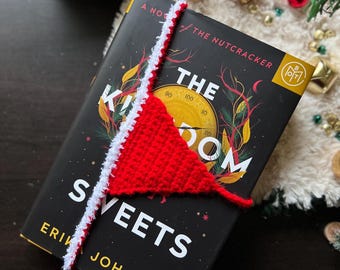 May include: A red and white crocheted bookmark rests on a book titled "The Kingdom of Sweets." The bookmark is shaped like a triangle with a white border. The book has a black cover with gold and red accents.