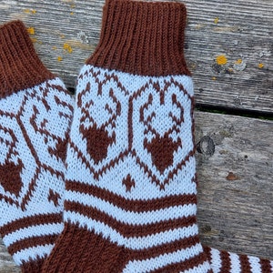 Può includere: Un paio di calzini di lana marroni e bianchi con un motivo a renna. I calzini sono su una superficie di legno.