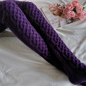 Peut inclure: Des bas en maille de couleur pourpre, à motifs torsadés et avec des pompons. Les bas sont posés sur une surface blanche.