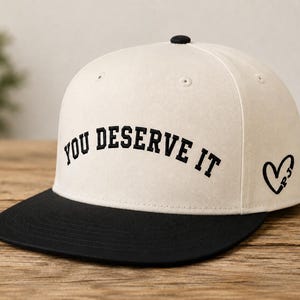 May include: A cream-colored baseball cap with a black flat brim and a black button on top. The front features the text YOU DESERVE IT embroidered in black block letters. A black heart outline with the letters PJ inside is on the side panel.