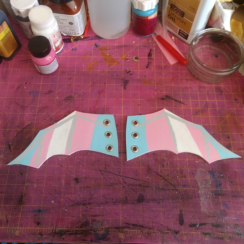 Pode incluir: Duas pe&ccedil;as pintadas &agrave; m&atilde;o em forma de asa. Cada asa apresenta um esquema de cores azul claro, rosa e branco com detalhes prateados. As asas t&ecirc;m ilh&oacute;s de metal para fixa&ccedil;&atilde;o, sugerindo um projeto de fantasia ou artesanato.