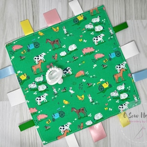 May include: A green baby sensory blanket with a farm animal print. The blanket has five fabric tags in different colors and a white teether attached to the center.