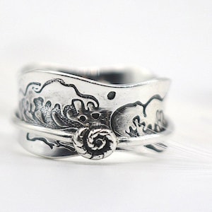 Könnte beinhalten: Ein silberfarbener Ring mit einem Wellenmuster und einem zentralen Spiraldetail. Der Ring weist aufwendige Schnitzereien von Meereswellen um das Band auf. Das Spiraldetail ist mittig auf dem Ring angeordnet.