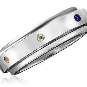 May include: A silver ring with three small gemstones in yellow, green, and blue. The ring has a polished surface and a contemporary design. The gemstones are set into the silver band. The ring is likely made of a metal such as stainless steel.