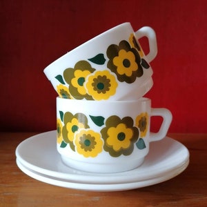 Puede incluir: Dos tazas de café de cerámica blanca con un patrón floral de flores amarillas y verdes. Las tazas están apiladas una encima de la otra y están sobre dos platillos blancos.