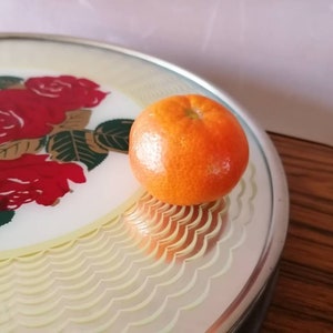 May include: A round serving tray with a floral rose design in red, gold, and green. An orange tangerine sits on the tray. The tray has a scalloped edge and a silver rim.
