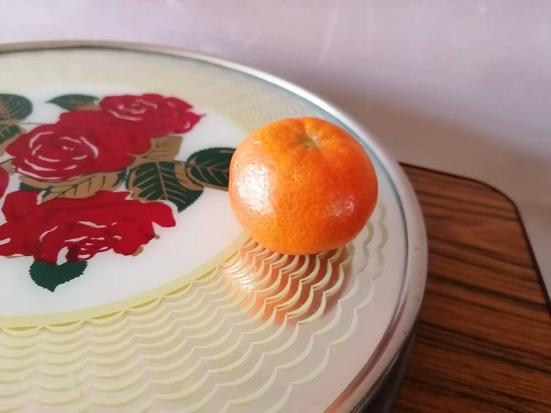 May include: A round serving tray with a floral rose design in red, gold, and green. An orange tangerine sits on the tray. The tray has a scalloped edge and a silver rim.