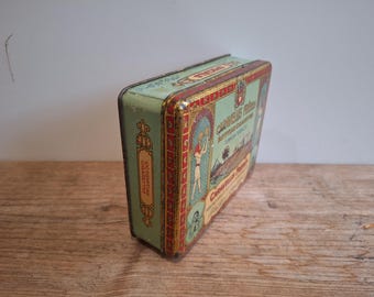 May include: A rectangular vintage metal cigarette tin featuring a light green background with red and gold decorative borders. The front displays an illustration of an Egyptian figure, palm trees, and the text Caravellas Freres Egyptian Cigarettes.