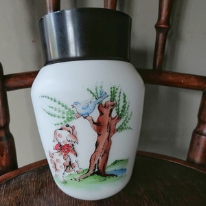 Może przedstawiać: Biały ceramiczny wazon z brązową pokrywką, z ręcznie malowanym wzorem przedstawiającym psa, ptaka i drzewo. Wazon ma fantazyjny i uroczy styl.
