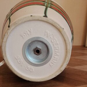 May include: Close-up of the bottom of a ceramic lamp base. The base is off-white with red and green stripes. The words "W. Germany" and "315-50" are stamped on the bottom. A metal plate and a power cord are also visible.