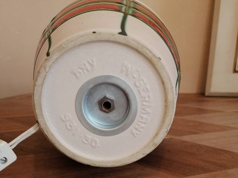 May include: Close-up of the bottom of a ceramic lamp base. The base is off-white with red and green stripes. The words "W. Germany" and "315-50" are stamped on the bottom. A metal plate and a power cord are also visible.