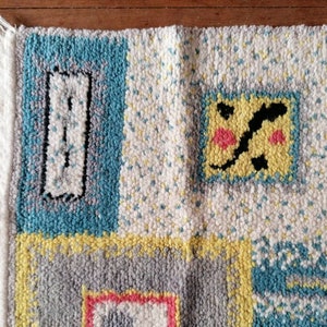 May include: A colorful rug with a geometric pattern. The rug is made of wool and has a fringe border. The colors in the rug are blue, yellow, gray, white, and pink.
