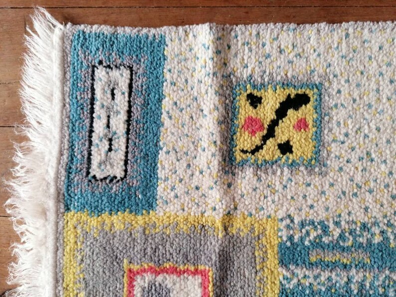 May include: A colorful rug with a geometric pattern. The rug is made of wool and has a fringe border. The colors in the rug are blue, yellow, gray, white, and pink.