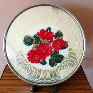 May include: A round, decorative plate with a silver rim. The plate features a central design of red roses with green leaves on a white background. The outer edge has a cream-colored, wavy pattern.