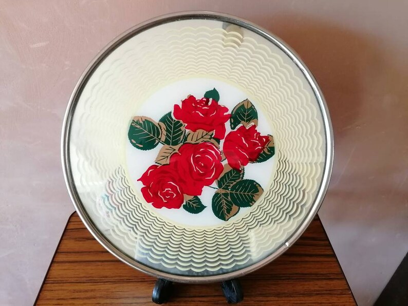 May include: A round, decorative plate with a silver rim. The plate features a central design of red roses with green leaves on a white background. The outer edge has a cream-colored, wavy pattern.