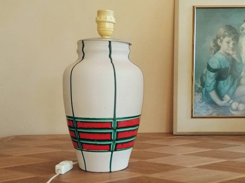 May include: White ceramic table lamp with a decorative design. The lamp features a cylindrical shape with a red and green geometric pattern around the middle. Vertical green lines accent the white body. The lamp has a standard light socket and a white cord.