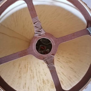 May include: A close-up view of the interior of a lampshade. The lampshade has a beige fabric stretched over a brown frame. The frame has a circular top and four brown supports that meet in the center. Metal clips and tape secure the frame.