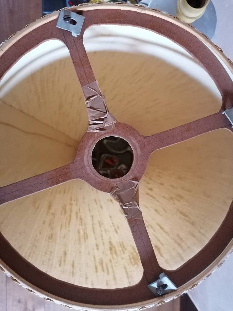 May include: A close-up view of the interior of a lampshade. The lampshade has a beige fabric stretched over a brown frame. The frame has a circular top and four brown supports that meet in the center. Metal clips and tape secure the frame.