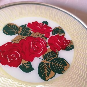 May include: A round, decorative plate with a floral design. The plate features a central image of red roses with green leaves and gold accents. The border has a scalloped pattern in a light yellow color, and the rim is silver.