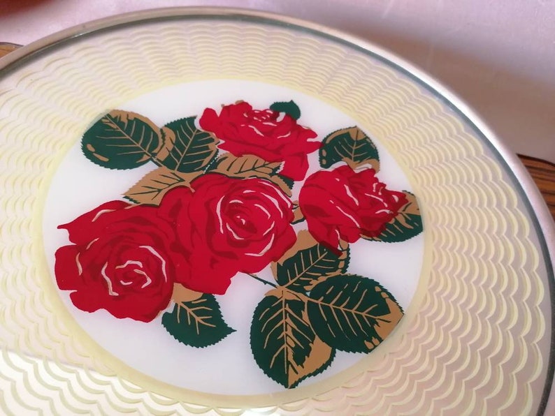 May include: A round, decorative plate with a floral design. The plate features a central image of red roses with green leaves and gold accents. The border has a scalloped pattern in a light yellow color, and the rim is silver.
