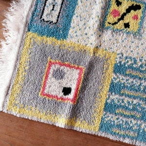 May include: A colorful rug with a patchwork design featuring geometric shapes and abstract patterns in shades of yellow, blue, gray, pink, and white. The rug has a white fringe border.