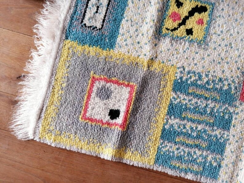 May include: A colorful rug with a patchwork design featuring geometric shapes and abstract patterns in shades of yellow, blue, gray, pink, and white. The rug has a white fringe border.