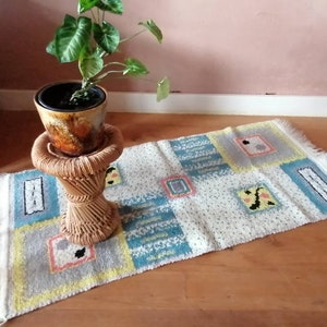 May include: A white rug with a geometric pattern in shades of blue, yellow, and pink. The rug has a fringe border and is placed on a wooden floor. A potted plant with green leaves sits on a wicker stool.